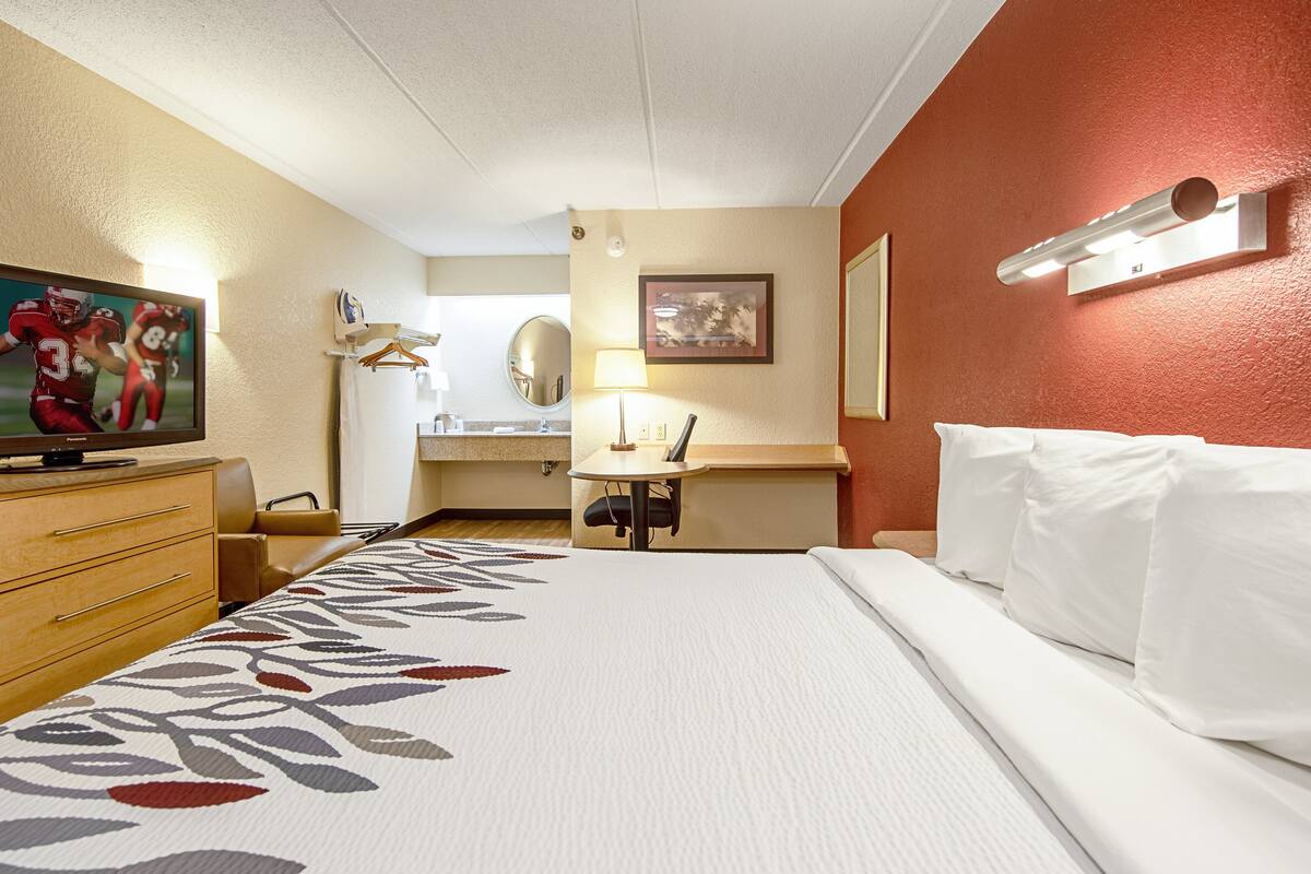 A hotel room with a bed, a desk, a chair, a television, a mirror, and a wall-mounted light fixture.