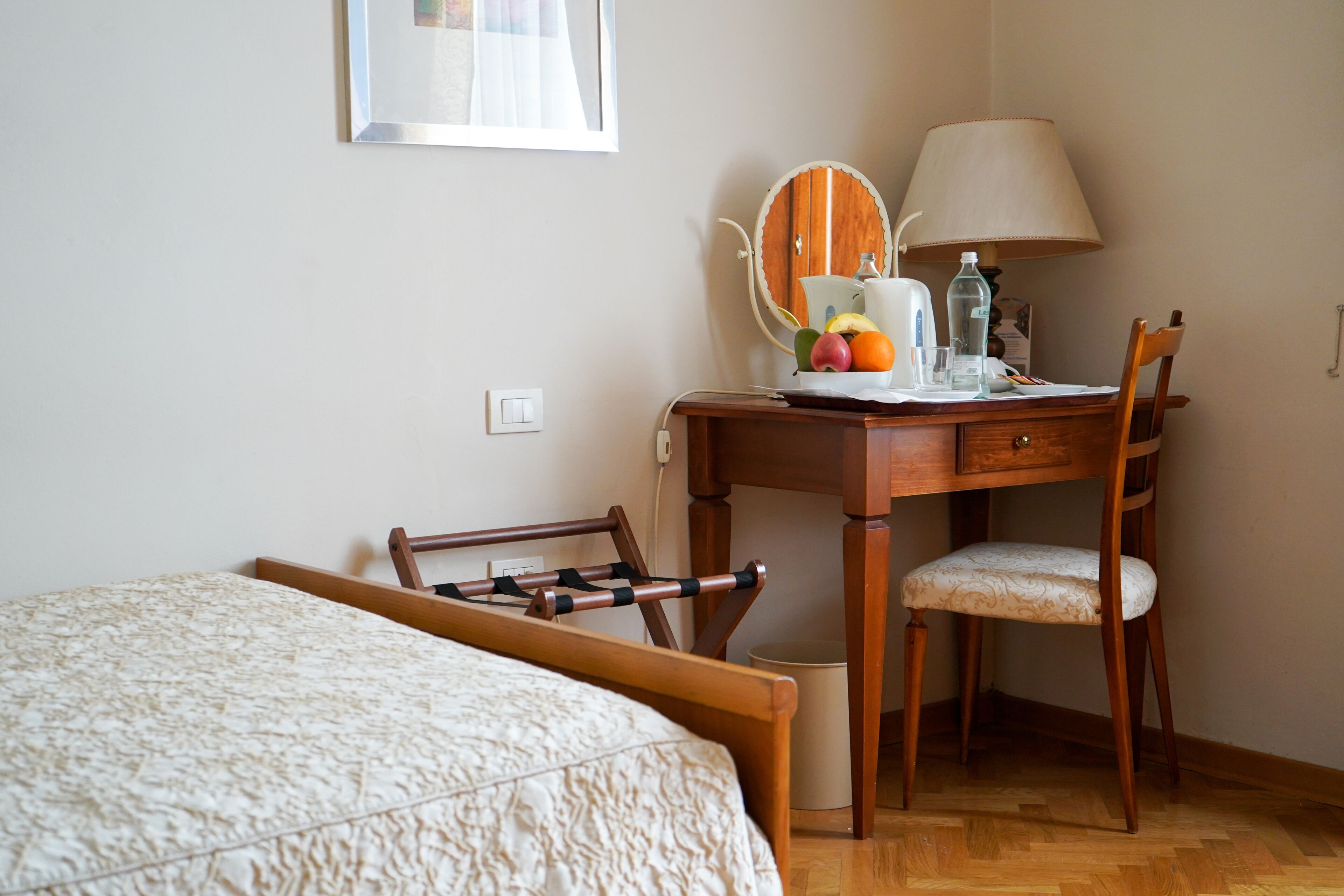 A bedroom with a bed, a wooden desk with a chair, a mirror, a lamp, and a small table with fruits and a water bottle.