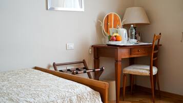 A bedroom with a bed, a wooden desk with a chair, a mirror, a lamp, and a small table with fruits and a water bottle.