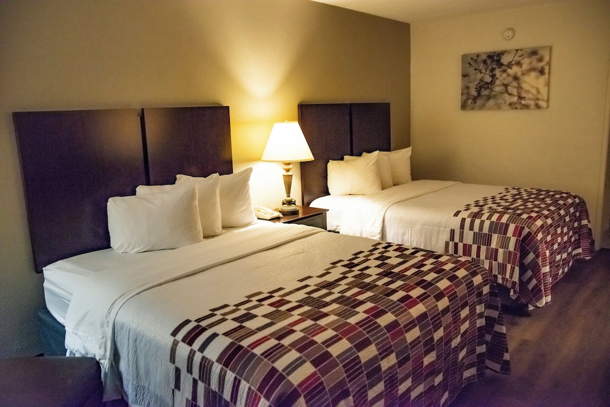 A hotel room with two beds, a nightstand with a lamp, and a wall-mounted artwork.