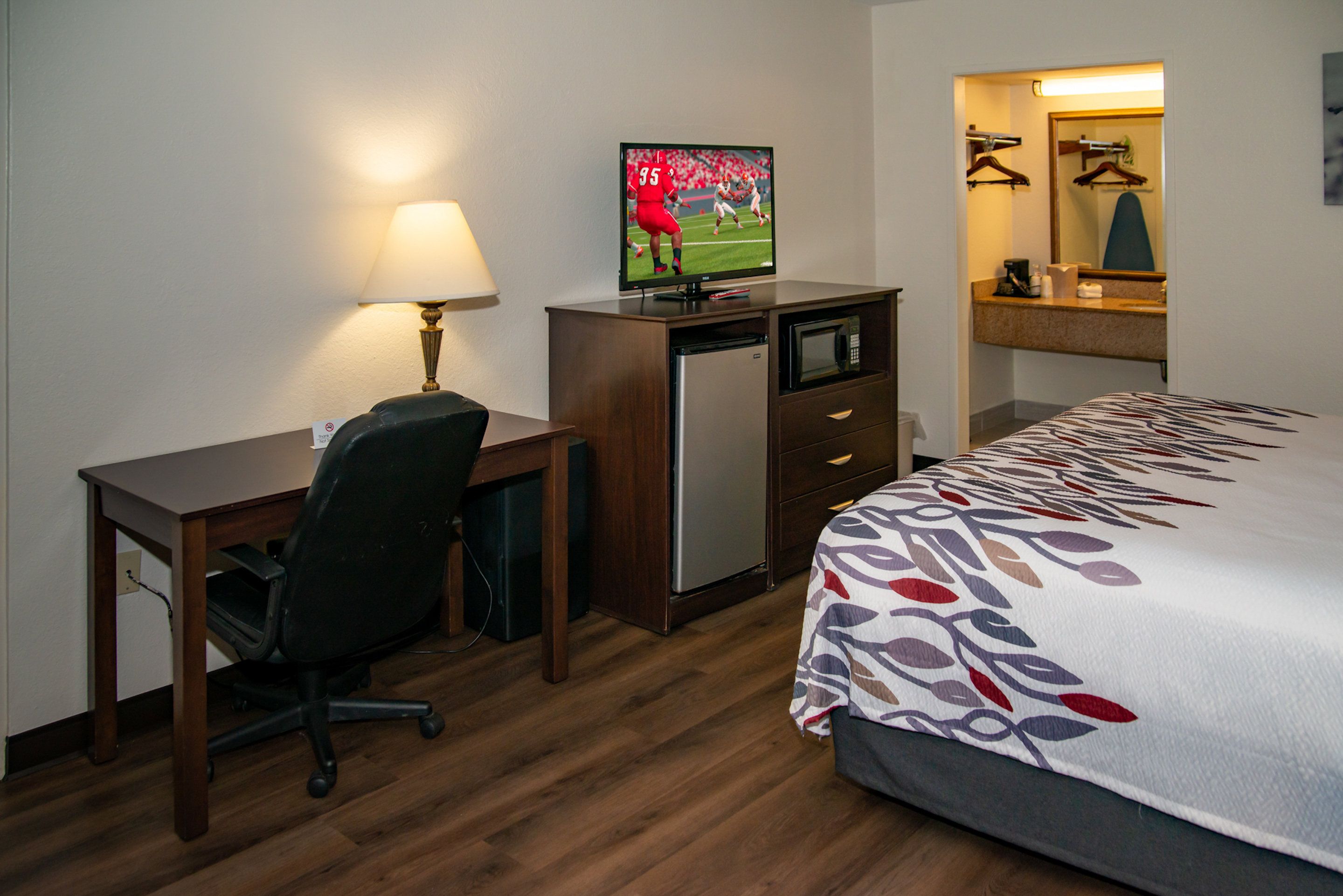 A hotel room with a bed, a desk, a chair, a television, a refrigerator, a microwave, and a mirror.