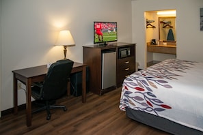 Habitación de hotel con cama, escritorio, silla, televisor, refrigerador y espejo.