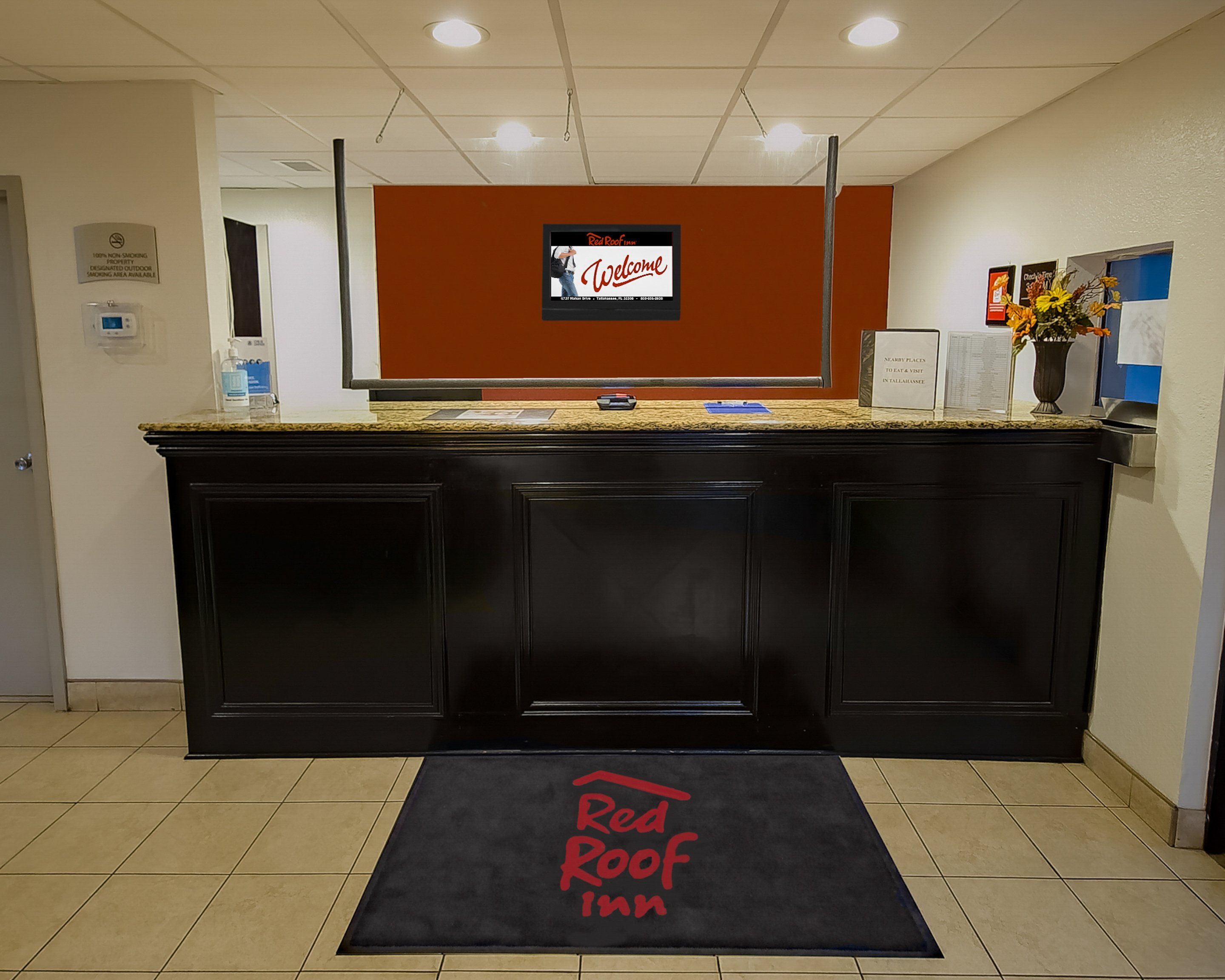 A reception desk with a digital screen displaying a welcome message, a Red Roof Inn sign, and a welcome mat at the entrance.