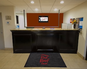 Een receptiebalie met een digitaal scherm waarop een welkomstboodschap staat, een Red Roof Inn bord en een welkomstmat bij de ingang.