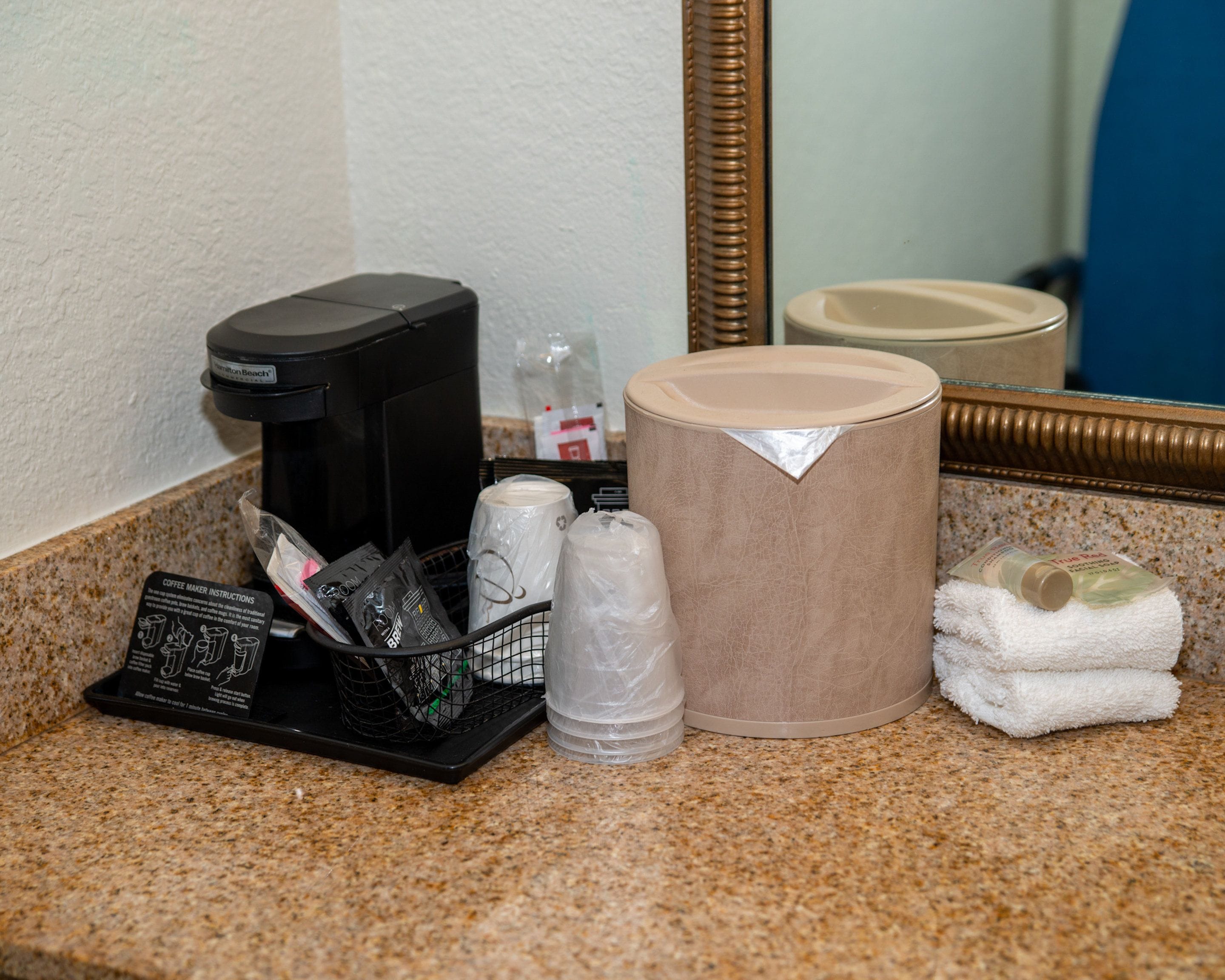 A coffee maker, a trash can, a basket with coffee pods, and a towel on a countertop.