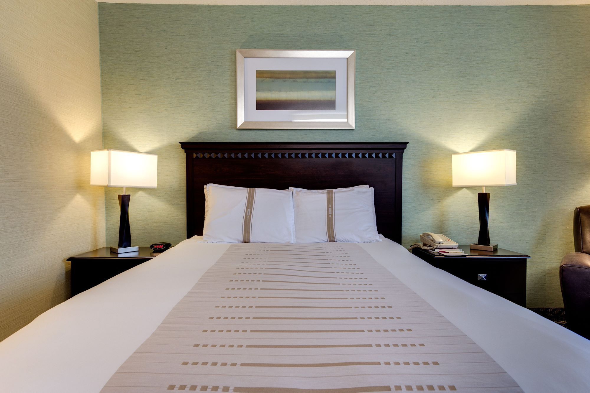 Una habitación de hotel con una cama grande, dos mesitas de noche con lámparas, un cuadro en la pared y un teléfono en una de las mesitas.