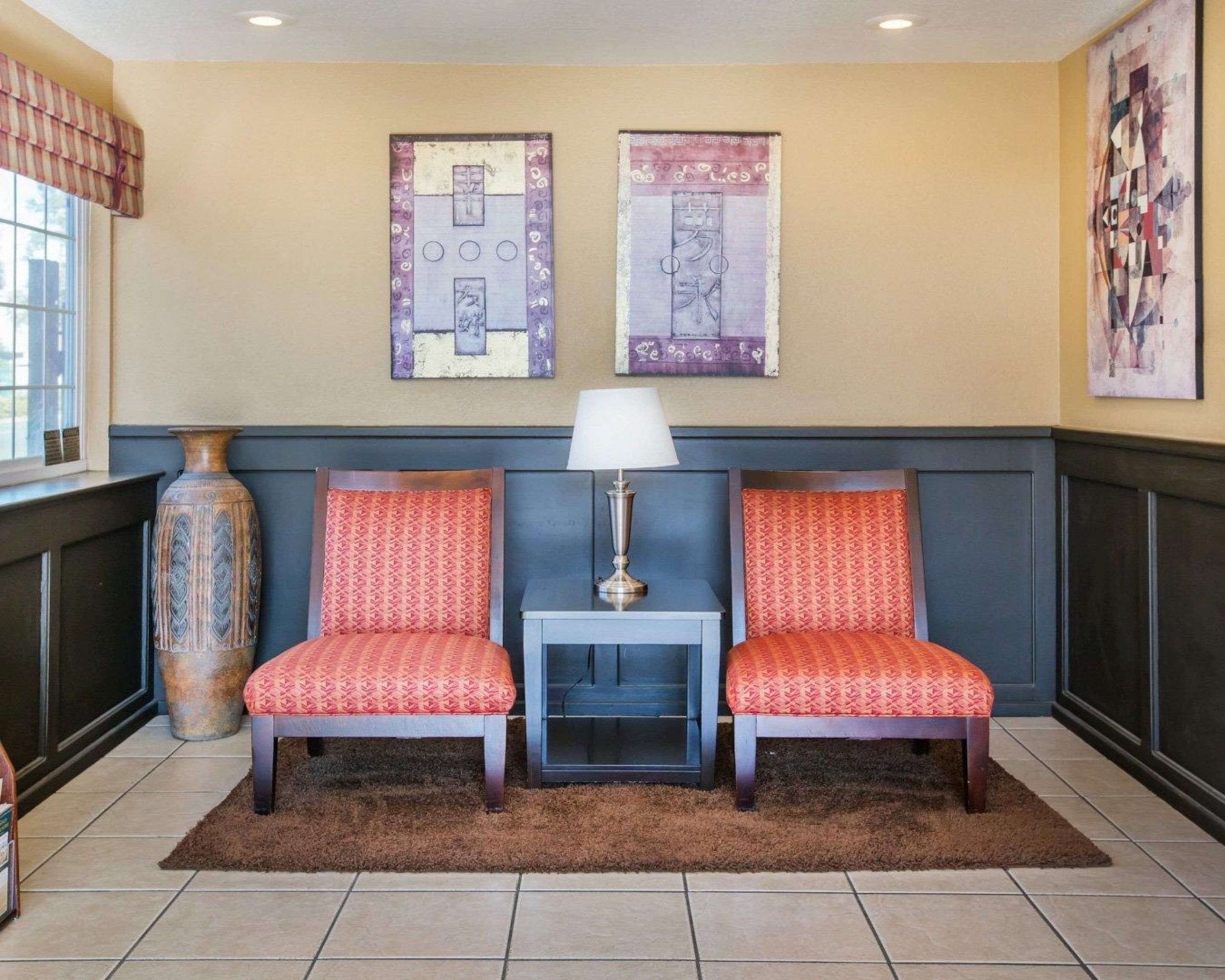 A hotel room with two red patterned chairs, a small table with a lamp, and two wall art pieces.