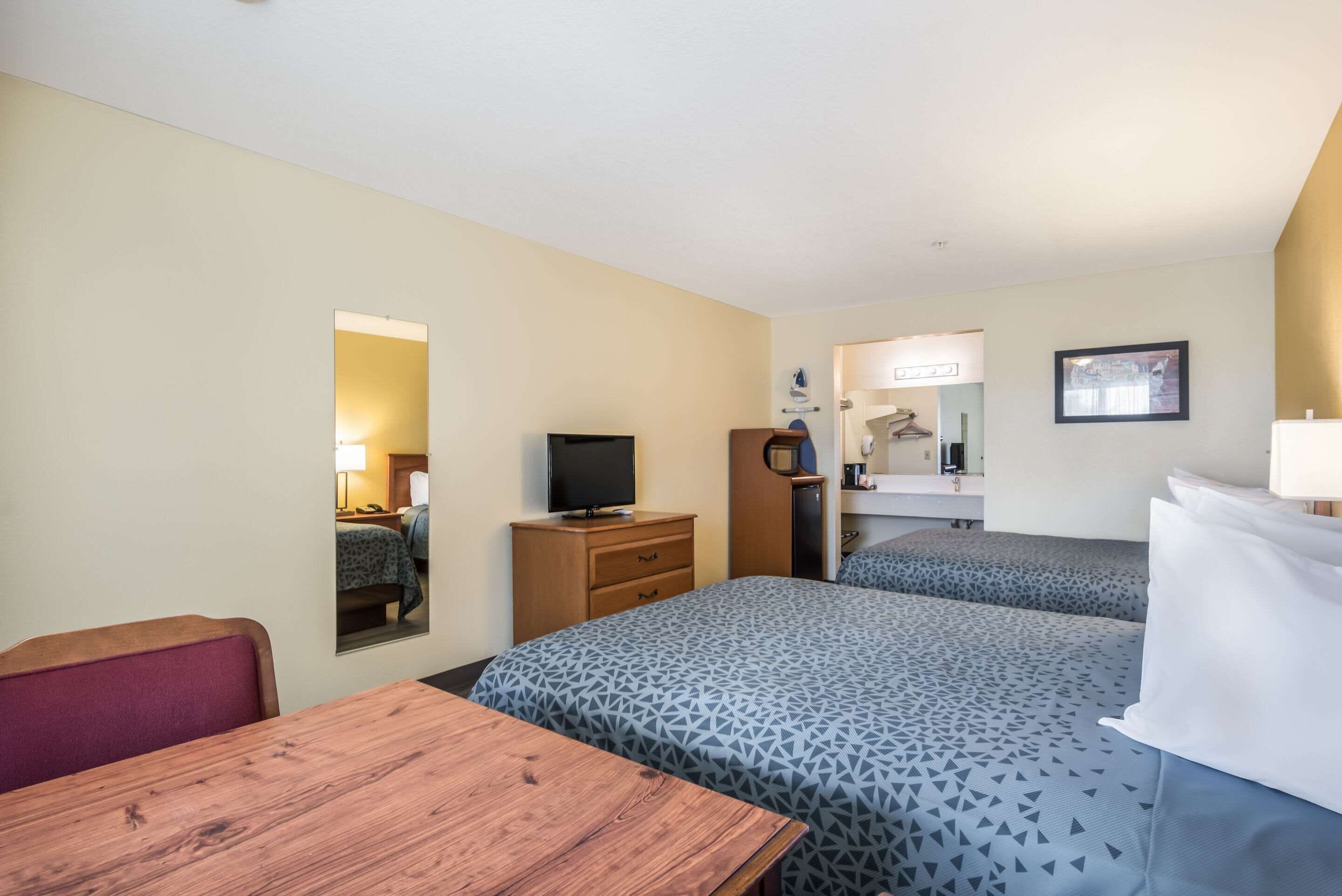 A hotel room with two beds, a wooden desk, a television, a mirror, and a small bathroom area.