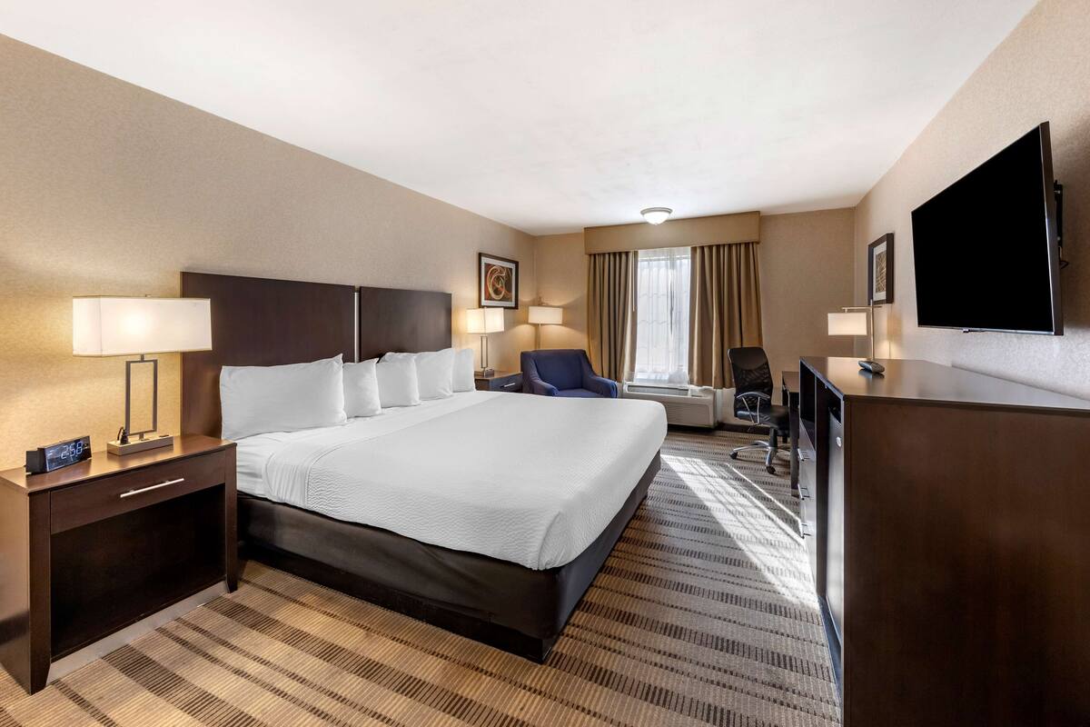 A hotel room with a large bed, bedside tables, a desk, a chair, a flat-screen TV, and a window with curtains.