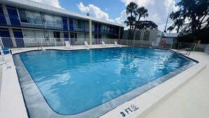 Una piscina rectangular con un letrero de 3 pies que indica 'no se permite bucear', rodeada por una terraza blanca y contigua a un edificio con barandillas azules y un balcón cubierto.