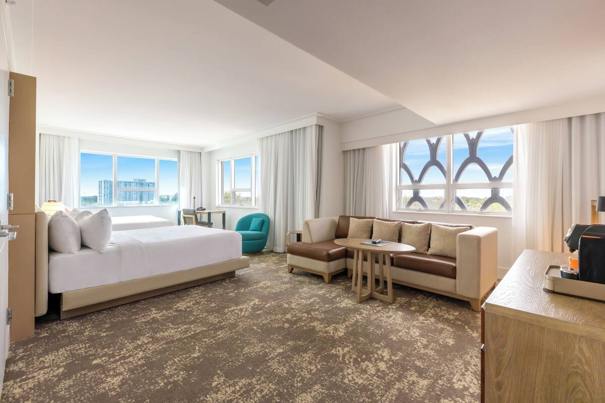 Habitación de hotel con una cama grande, un sofá, una mesita y un escritorio. Amplias ventanas con cortinas y vistas a la ciudad.