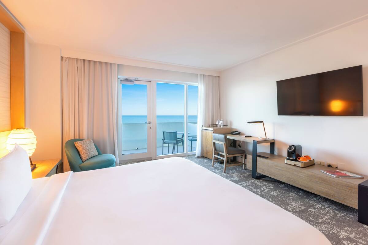 Habitación de hotel con una cama grande, un escritorio con silla, televisor montado en la pared y vista al mar por la ventana.
