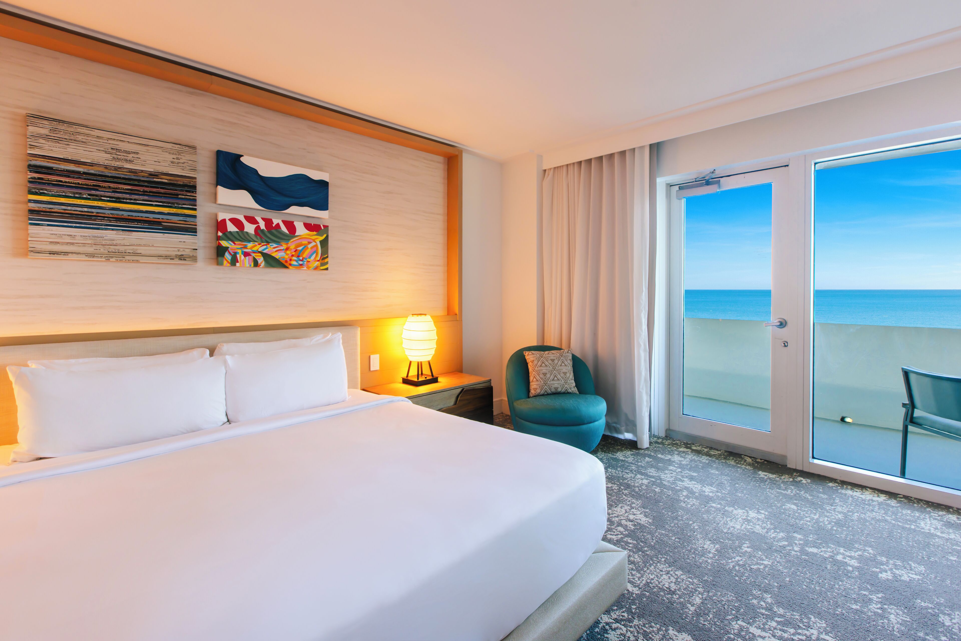 Camera d'albergo con un letto grande, un comodino con una lampada, una sedia e una porta vetrata scorrevole che dà su un balcone con vista sull'oceano.