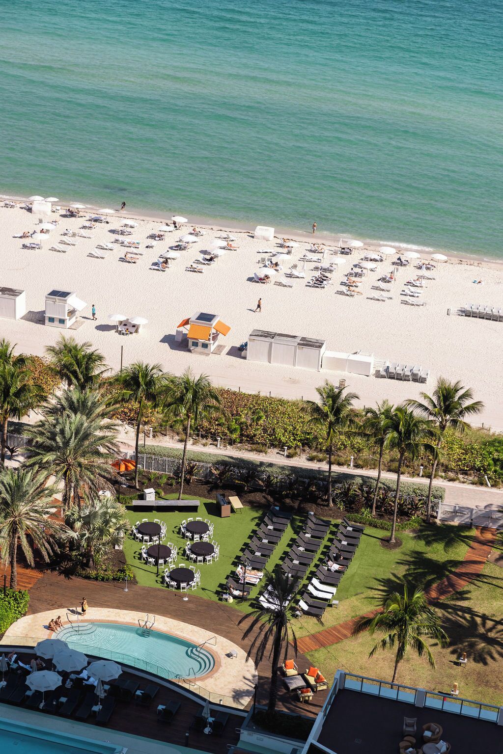 An aerial view of a beach resort with a swimming pool, sun loungers, and palm trees.