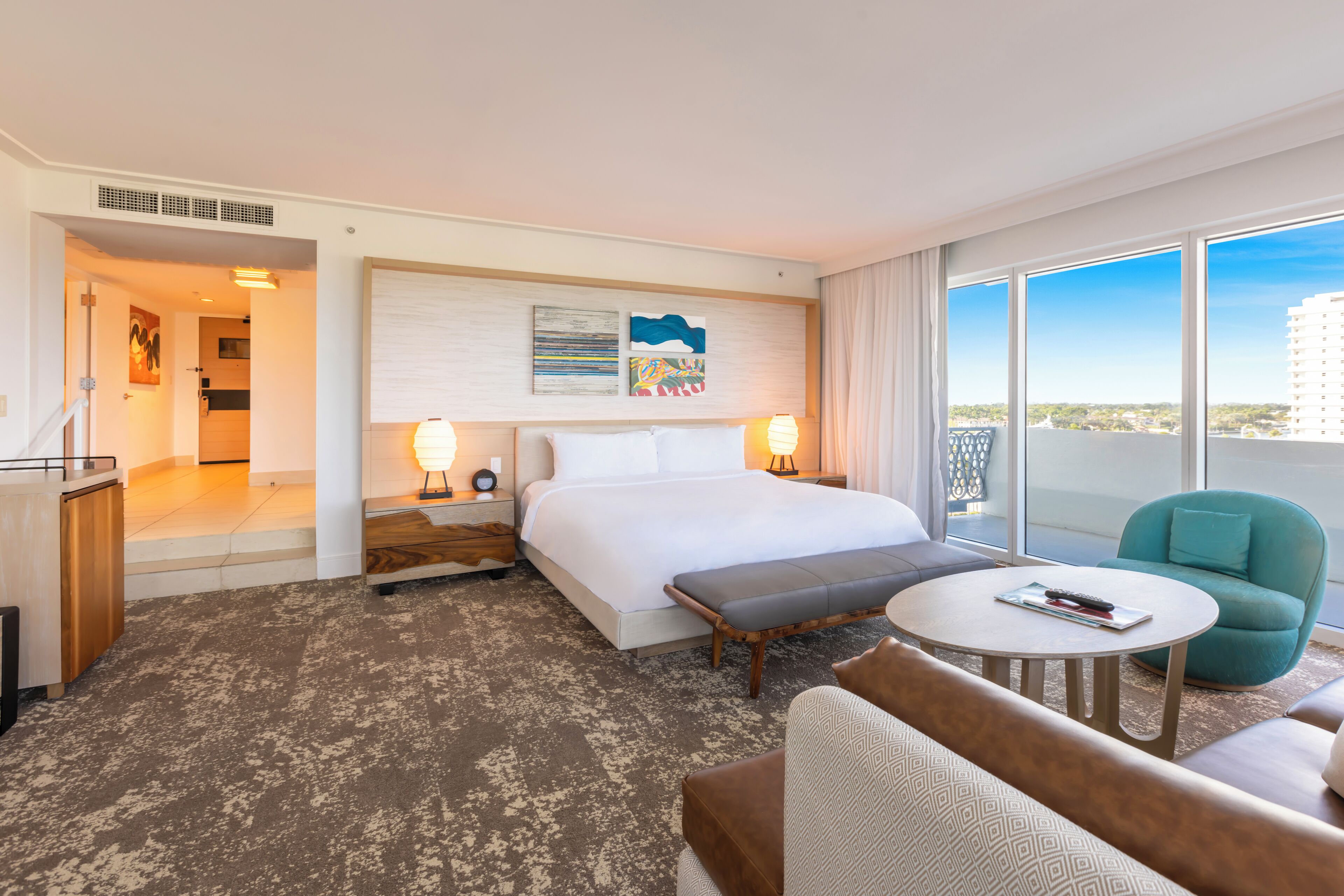 A hotel room with a bed, a desk, a round table, a sofa, and a large window offering a city view.