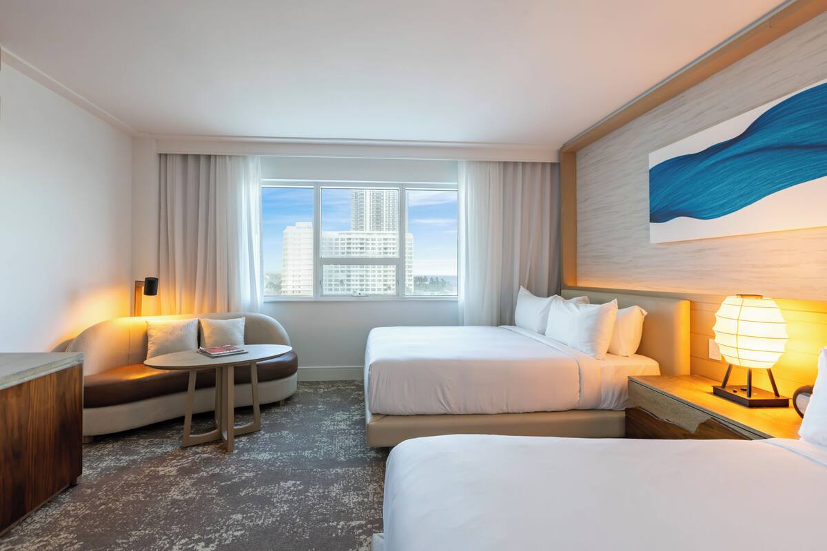 Habitación de hotel con dos camas, un sofá, una mesita, una lámpara y un ventanal con vistas a edificios.