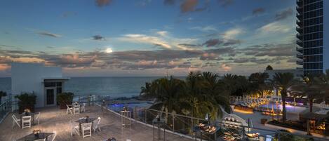 Una terraza en la azotea con mobiliario exterior, una piscina y vistas al océano al atardecer.