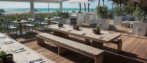 Een buitenterras met houten tafels en banken, omringd door potplanten en met uitzicht op een strand.