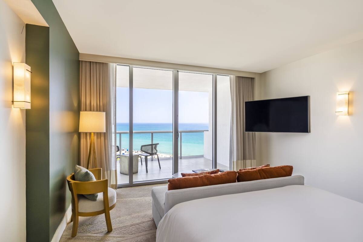 Una habitación de hotel con una cama grande, un televisor montado en la pared, un escritorio con una silla y un balcón con vista al océano.