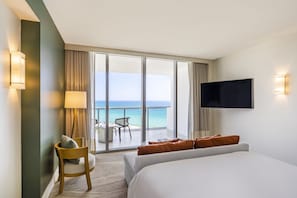 A hotel room with a large bed, a TV mounted on the wall, a desk with a chair, and a balcony with a view of the ocean.