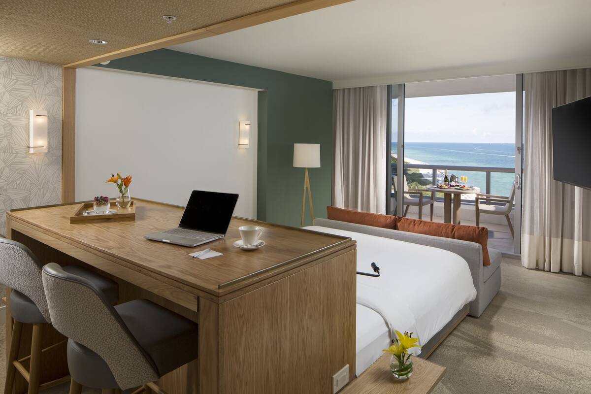 Habitación de hotel con un escritorio de madera, una laptop, una taza y un florero con flores. Se ve una cama con una colcha blanca y una vista al océano a través de amplias ventanas.