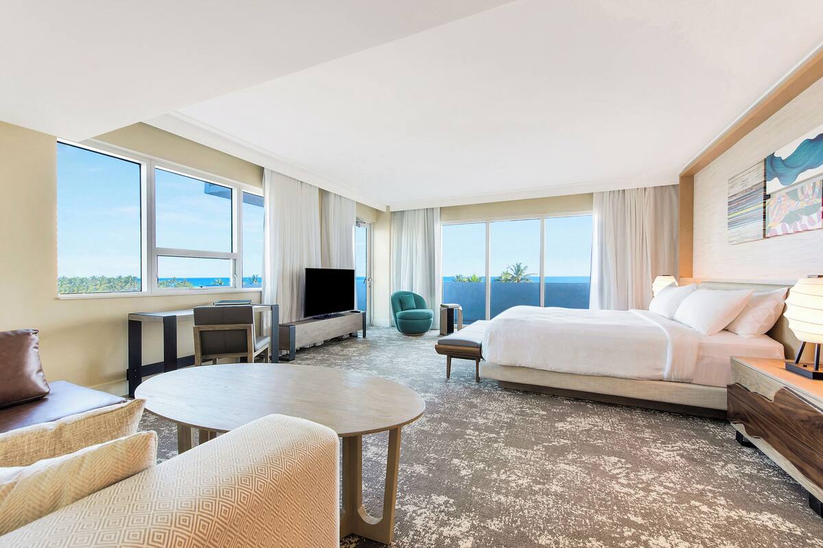 Habitación de hotel con cama, televisor, escritorio, silla y ventanas amplias con vista al océano.