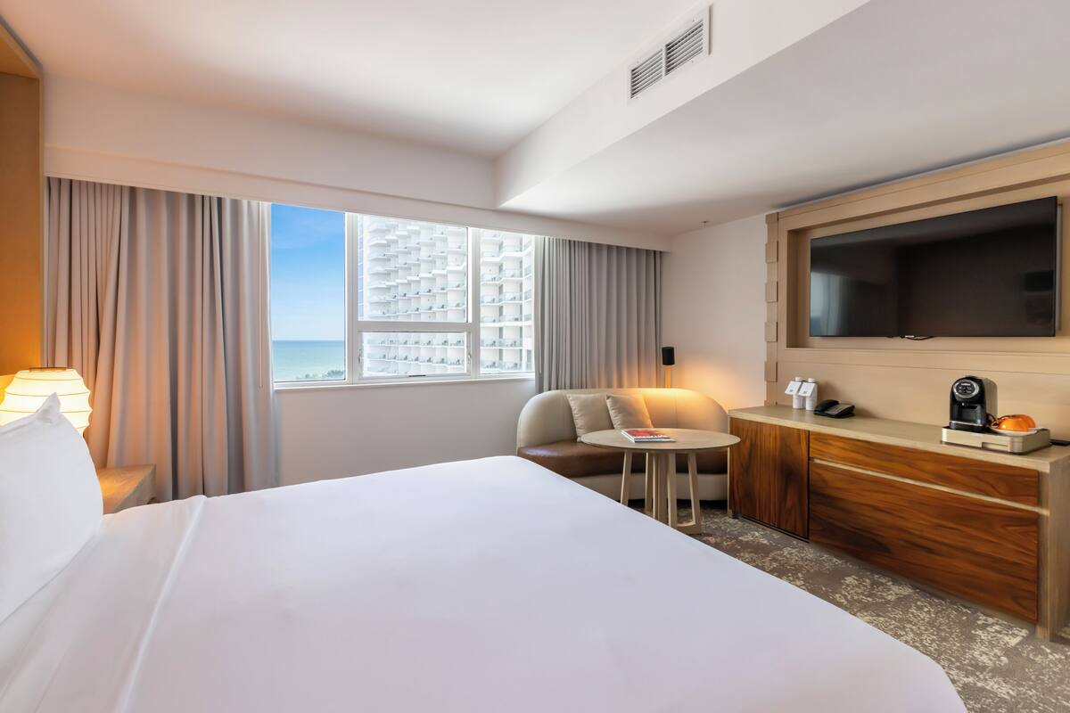Habitación de hotel con una cama grande, un televisor montado en la pared, una mesita, una silla y vistas a la ciudad desde la ventana.