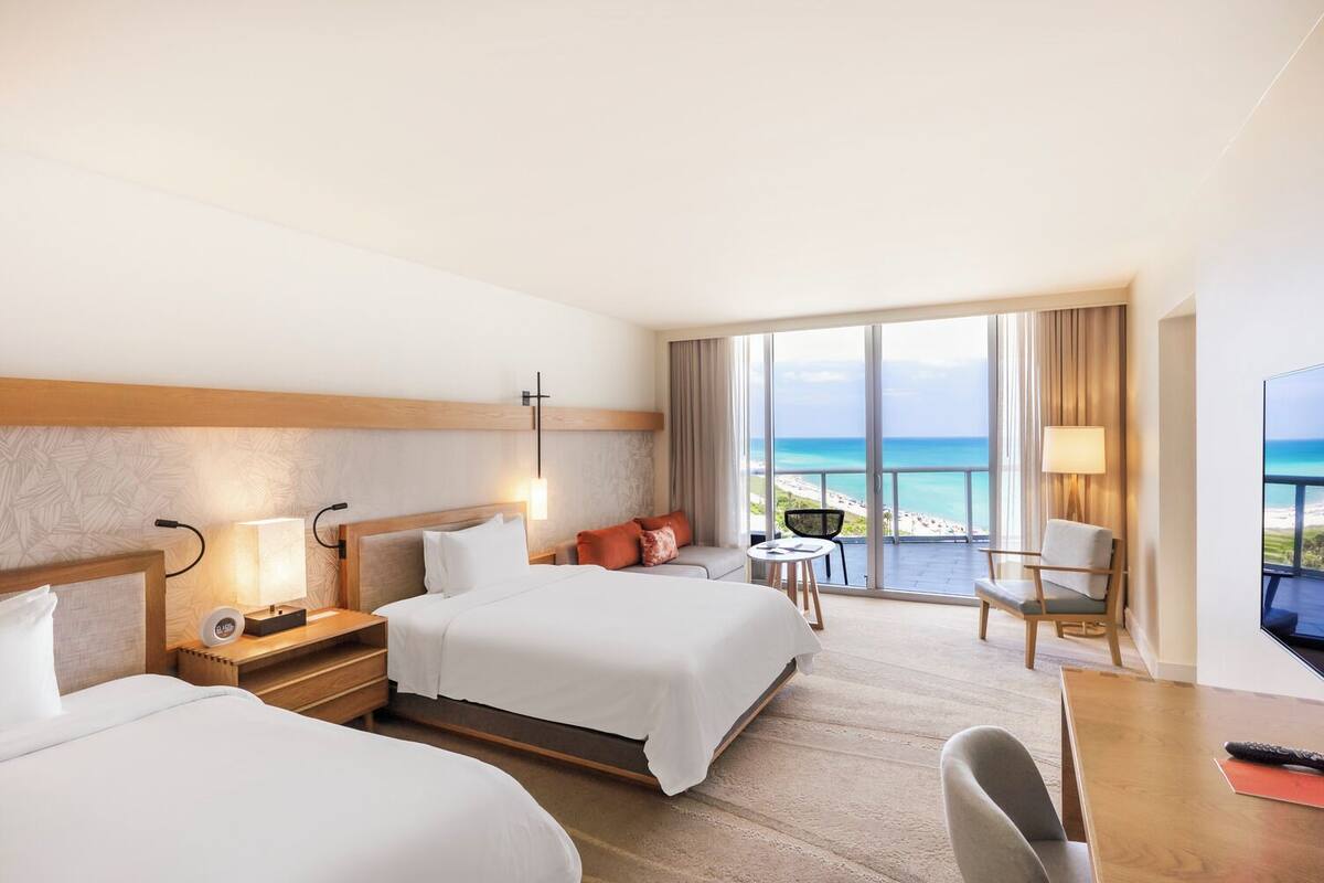 Habitación de hotel con dos camas, un escritorio, una silla, un televisor y un balcón con vistas a la playa.