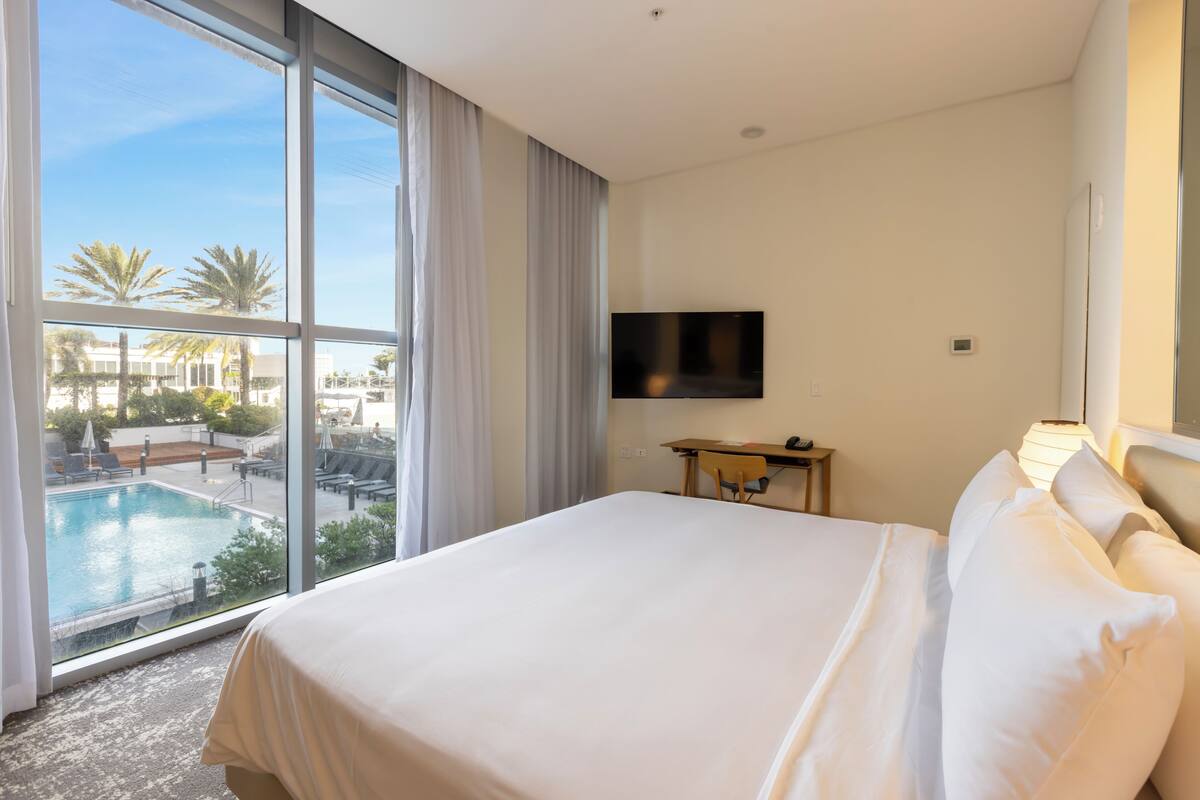 Habitación de hotel con una cama grande, un televisor montado en la pared y vista a un área de piscina al aire libre.