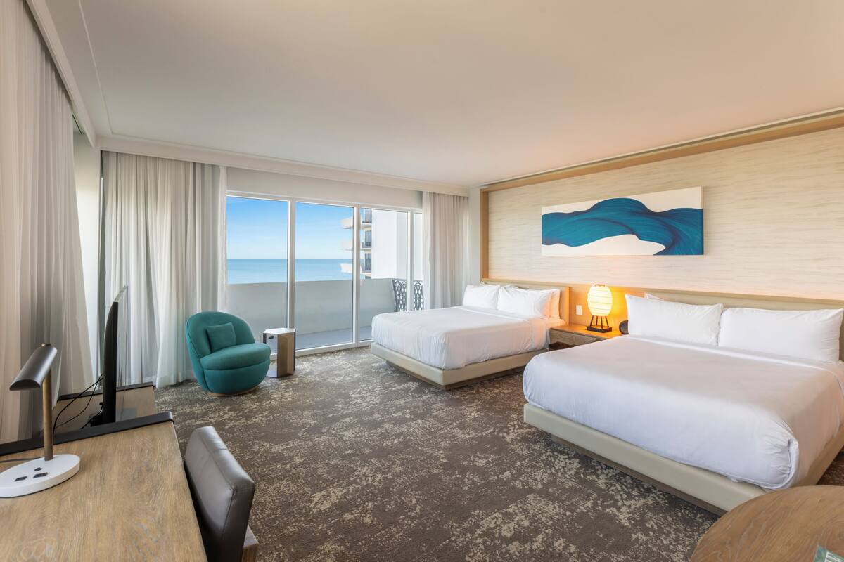 Habitación de hotel con dos camas, un escritorio con computadora, una silla, un televisor, una lámpara y un cuadro en la pared.