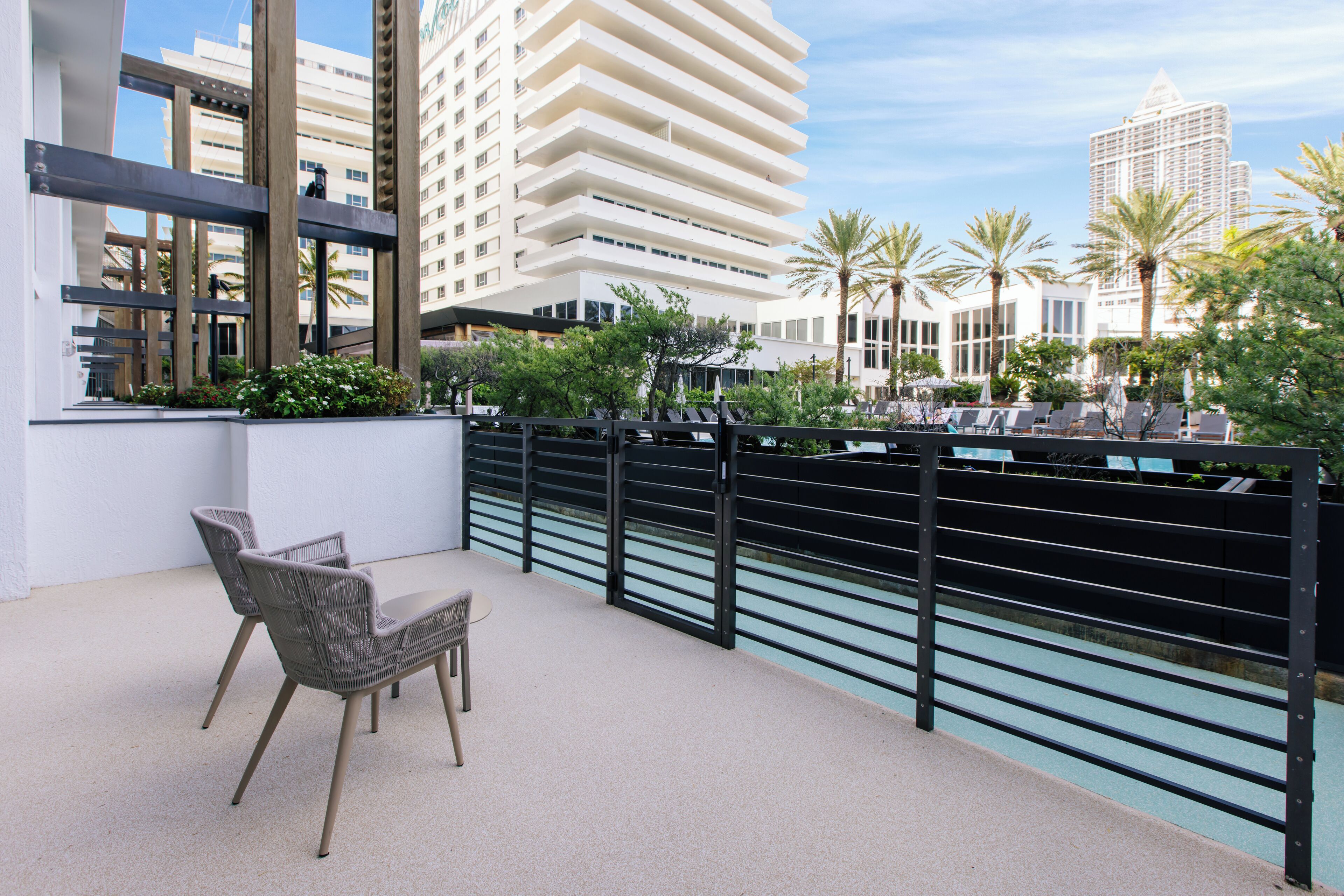 A balcony with wicker chairs and a table, overlooking a pool and palm trees, with modern buildings in the background.