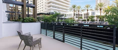 Un balcón con sillas de mimbre y una mesa, con vistas a una piscina y palmeras, y edificios modernos al fondo.