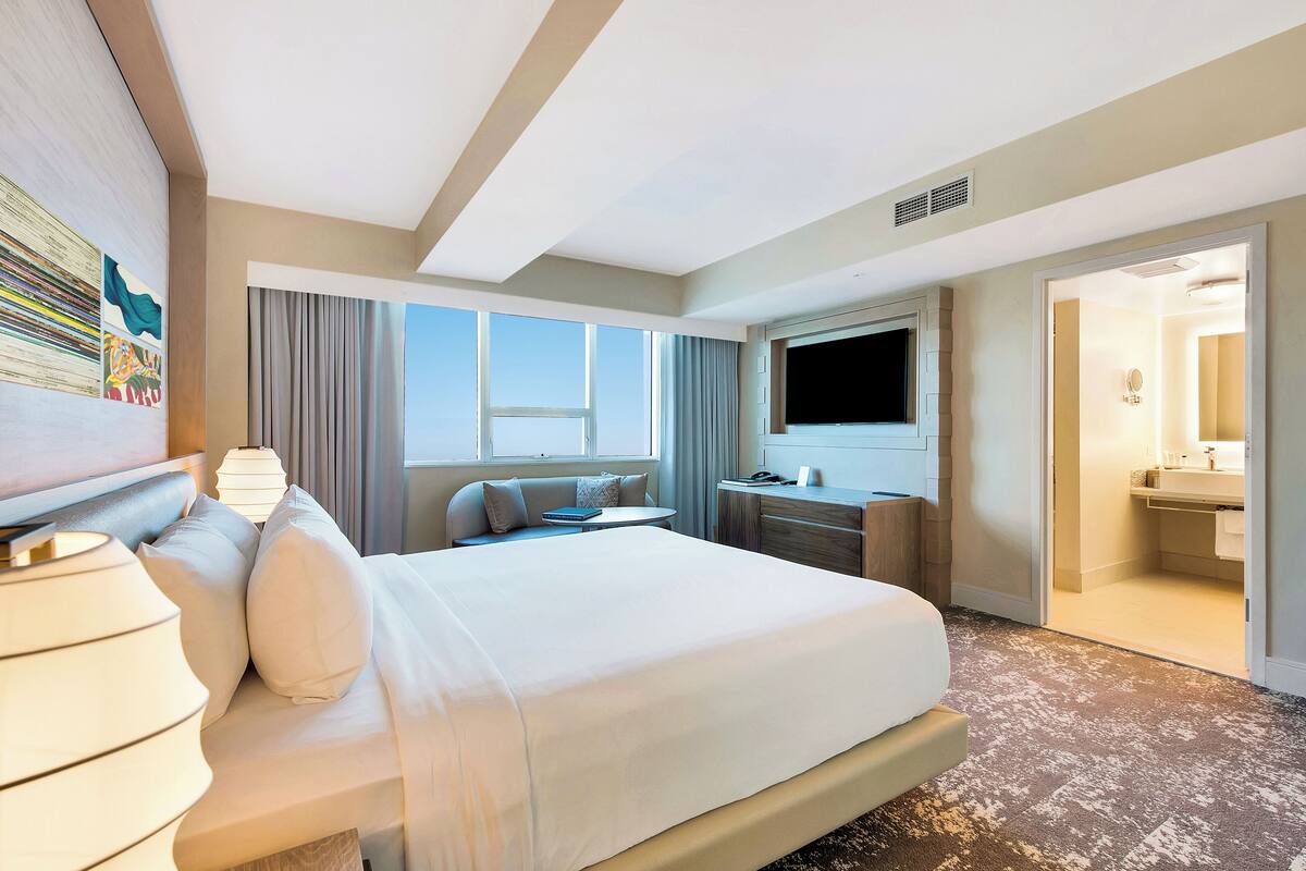 Habitación de hotel con una cama grande, un escritorio, un televisor y un baño visible a través de una puerta abierta.