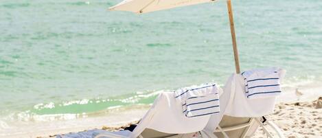 Two beach chairs under a white umbrella on a sandy beach with a calm sea in the background.