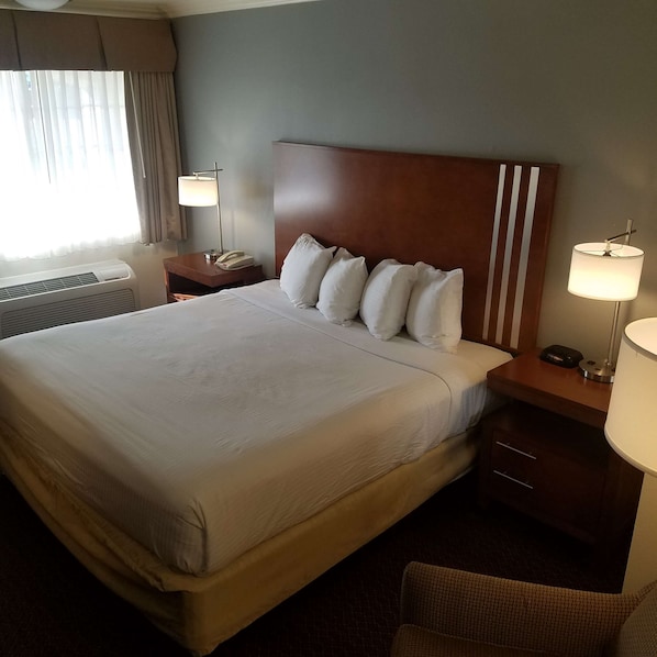 Una habitación de hotel con una cama grande, un cabecero de madera, dos mesitas de noche con lámparas, una silla y una ventana con cortinas.