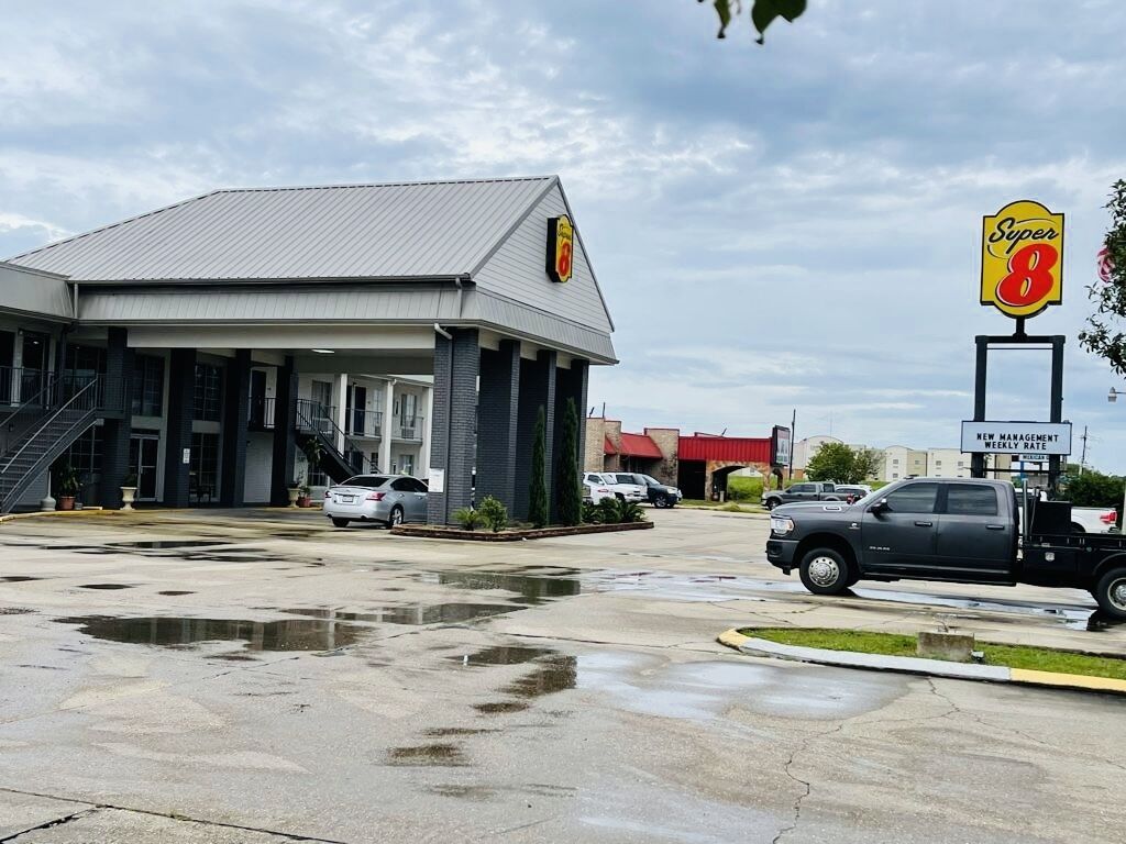 A Super 8 motel with a large parking lot, a covered entrance, and a sign indicating a new management weekly rate.