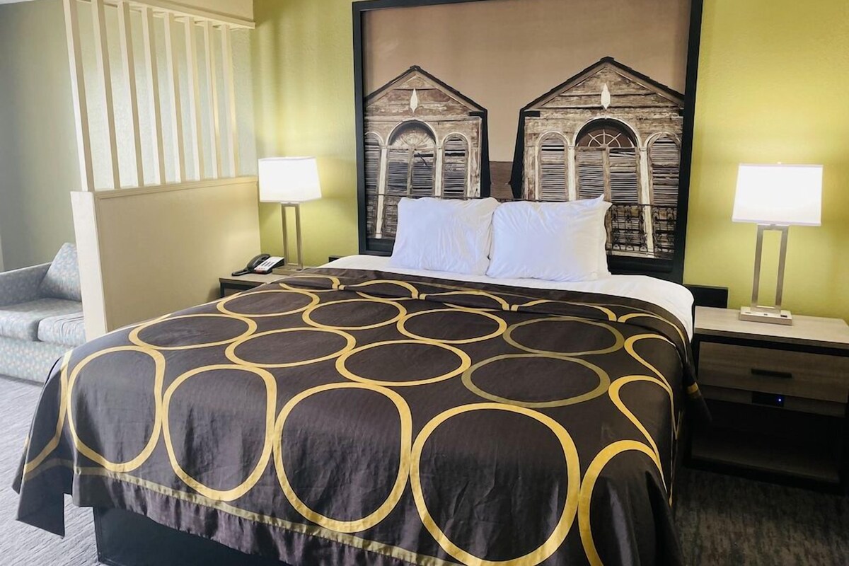 A hotel room with a large bed, two bedside tables, a nightstand, a wall-mounted lamp, and a painting of old buildings.