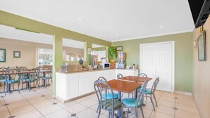 Un espace repas avec une table centrale et des chaises, un comptoir de cuisine avec des appareils électroménagers, et une porte à l’arrière-plan.