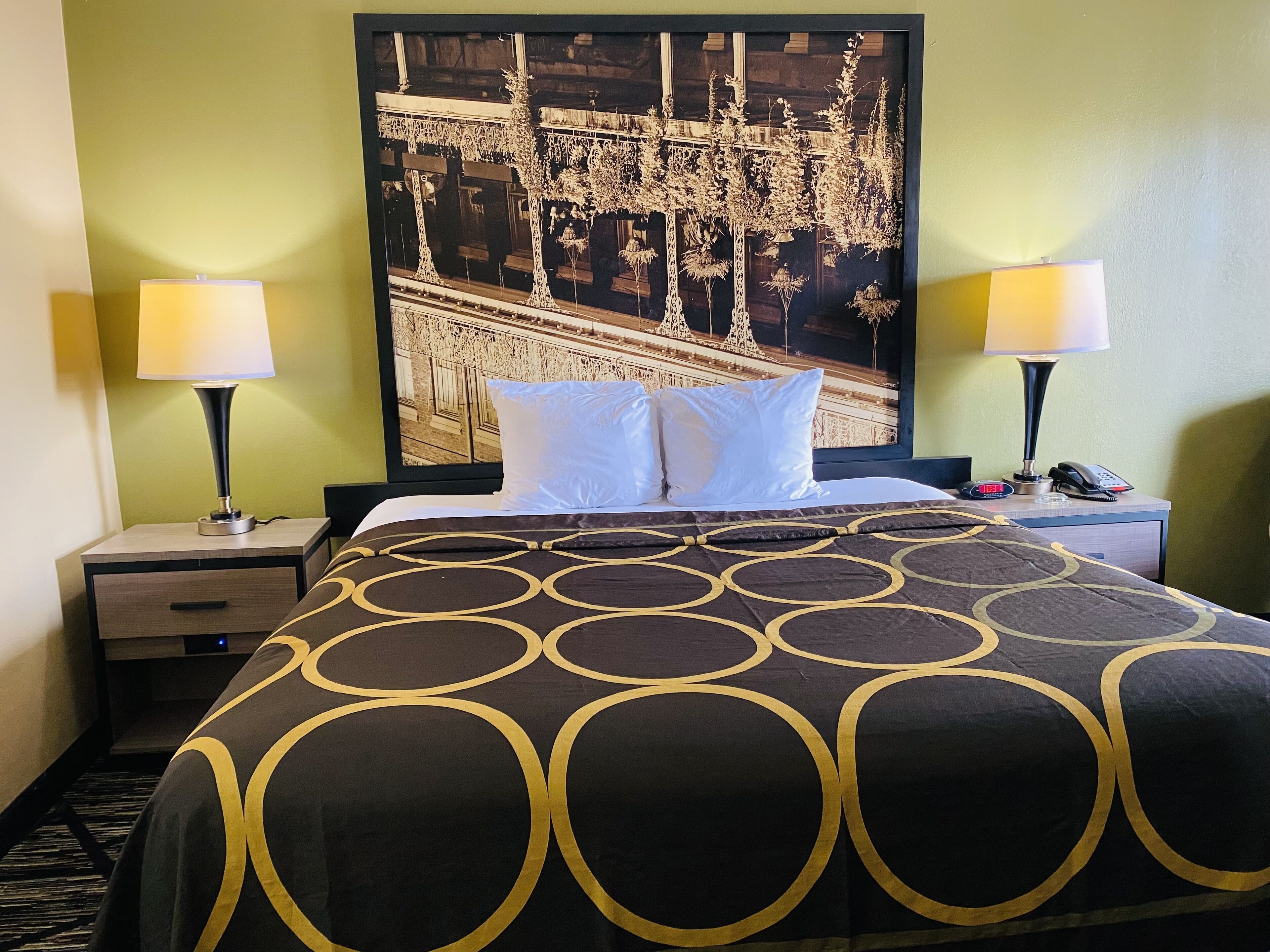 Habitación de hotel con cama, dos lámparas de noche, mesita de noche con teléfono y reloj, y un cuadro grande en la pared.