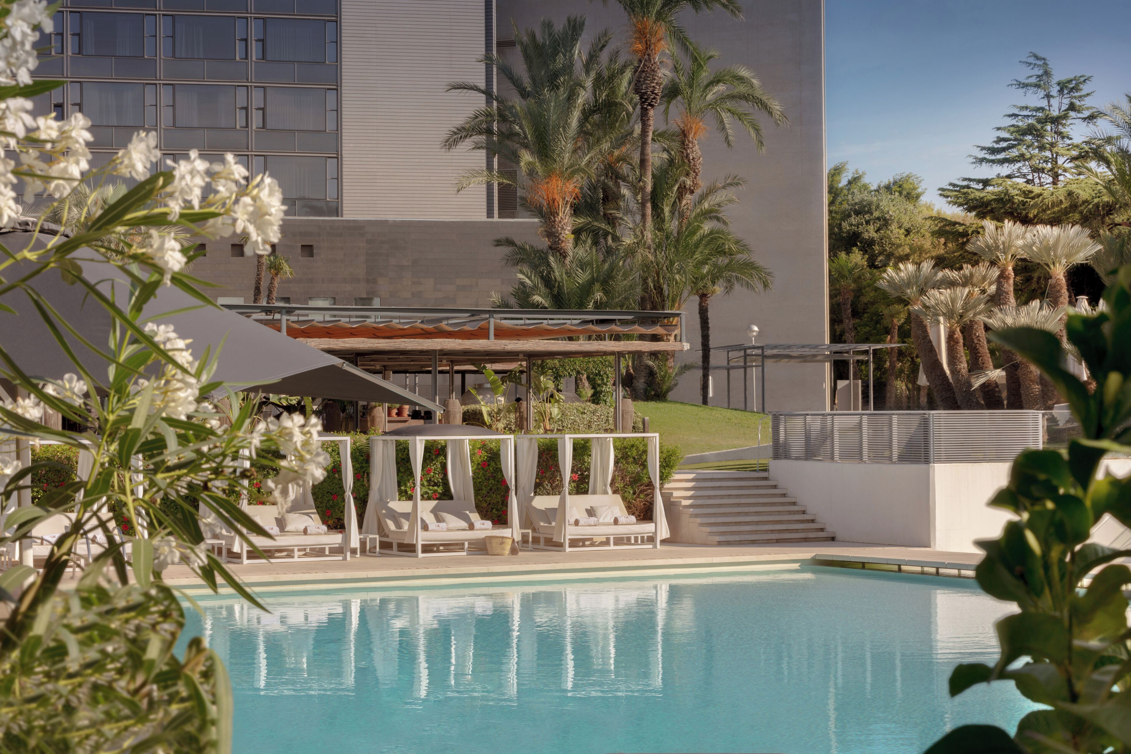 Zona de piscina con sillones de descanso, un espacio de estar cubierto y un edificio de diseño moderno al fondo.