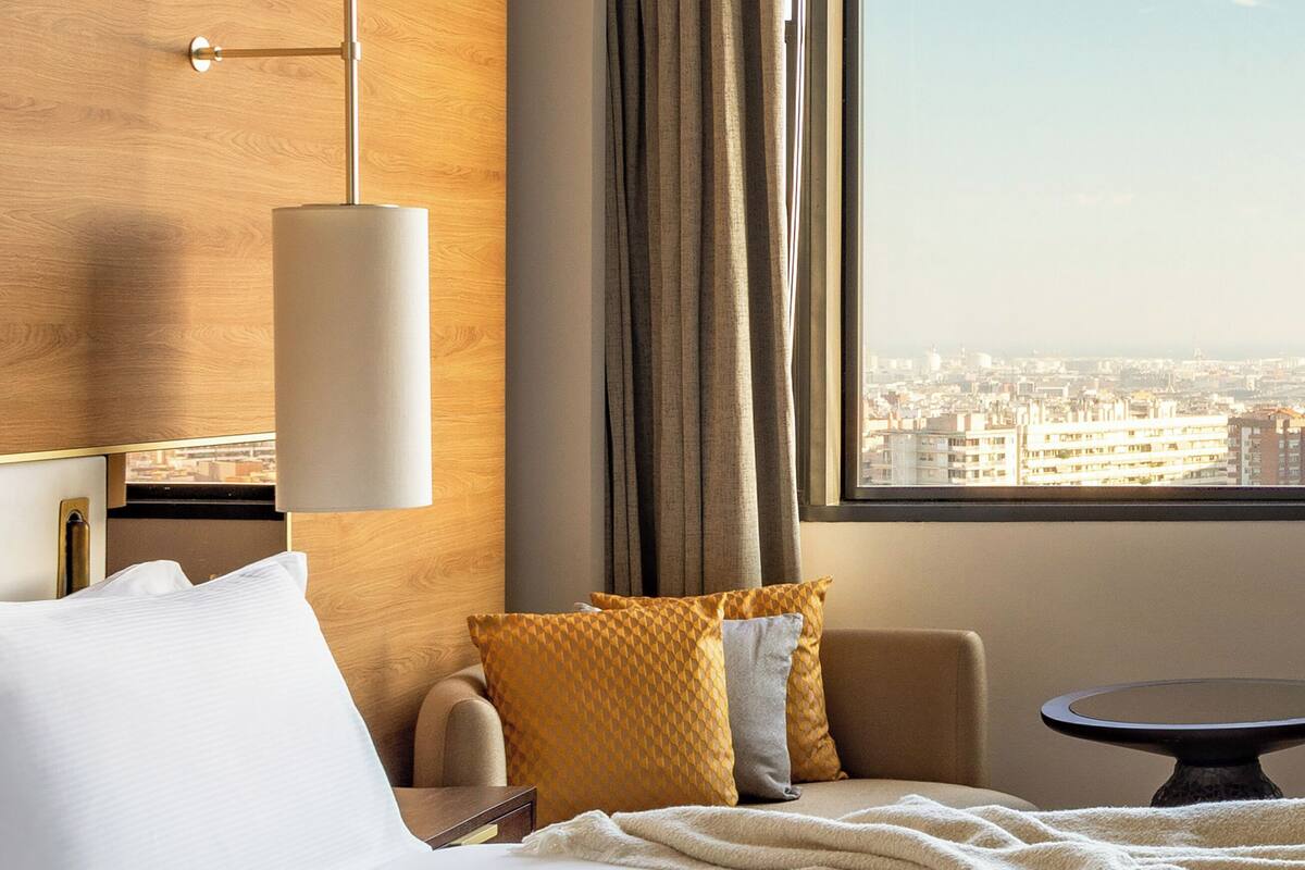 Una habitación de hotel con una cama, una mesita de noche, una lámpara, una silla, una mesa redonda y vistas de la ciudad a través de una ventana.