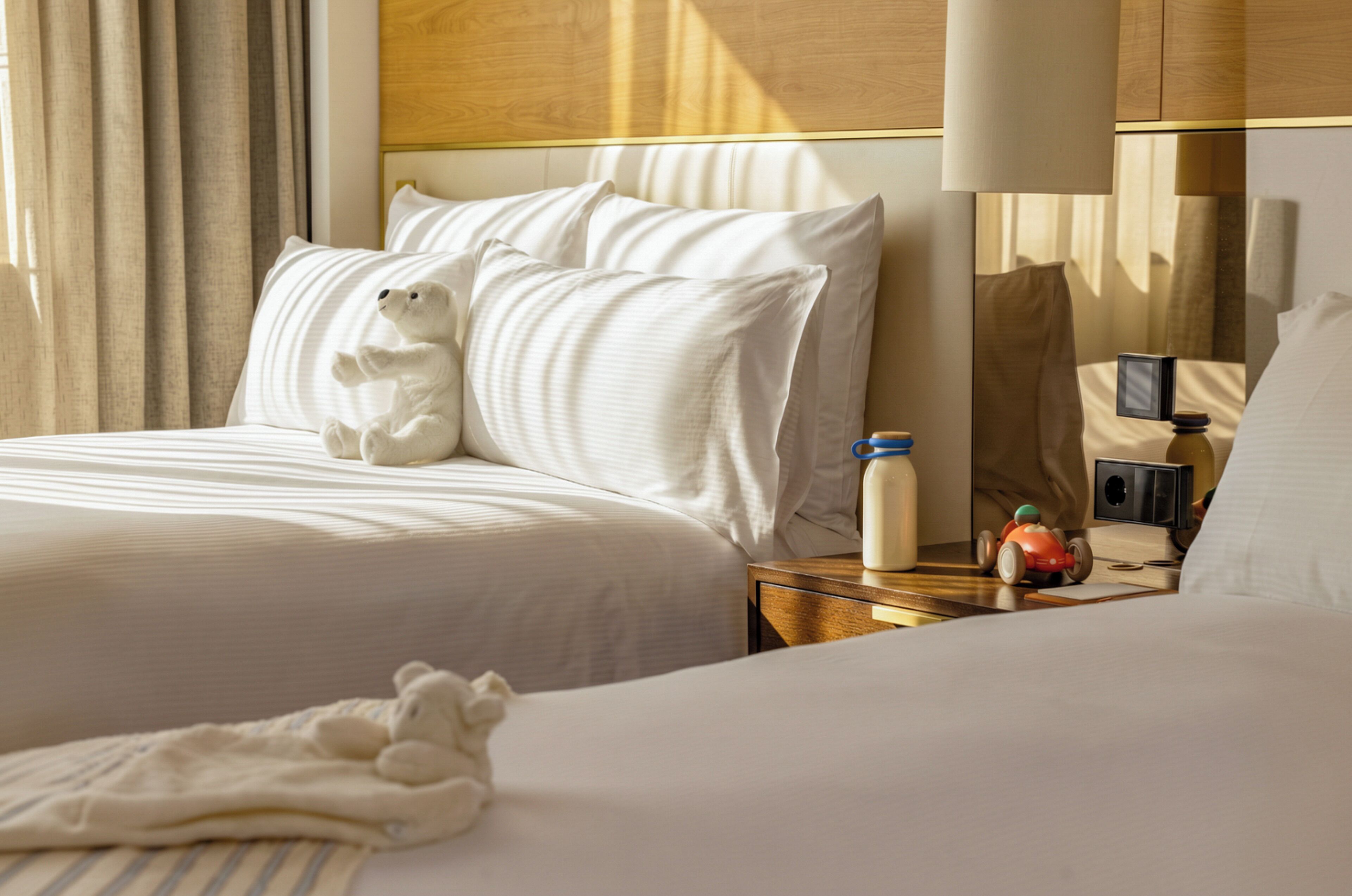 Ein Hotelzimmer mit einem Bett, Kissen, einem Teddybär, einem Nachttisch mit einer Flasche, einer Fernbedienung und einem Spielzeugauto.