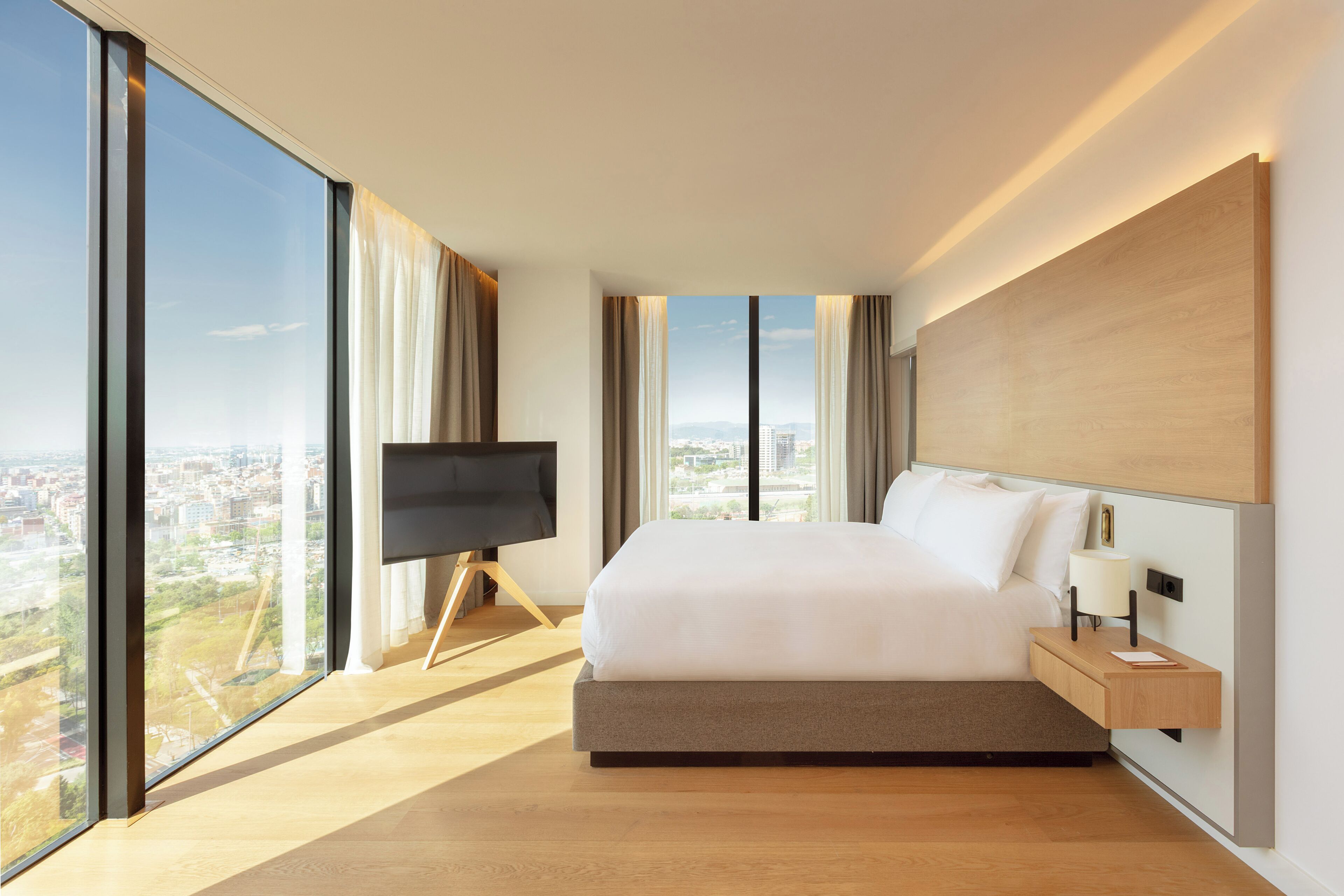 Una habitación de hotel moderna con una cama grande, un televisor en un mueble bajo, una mesita de noche con lámpara y vistas a la ciudad a través de amplios ventanales.