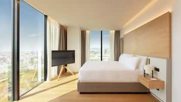 Una habitación de hotel moderna con una cama grande, un televisor en un mueble bajo, una mesita de noche con lámpara y vistas a la ciudad a través de amplios ventanales.
