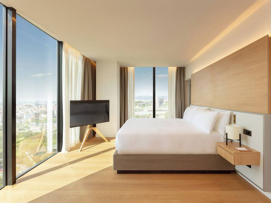 Una habitación de hotel moderna con una cama grande, un televisor en un mueble bajo, una mesita de noche con lámpara y vistas a la ciudad a través de amplios ventanales.