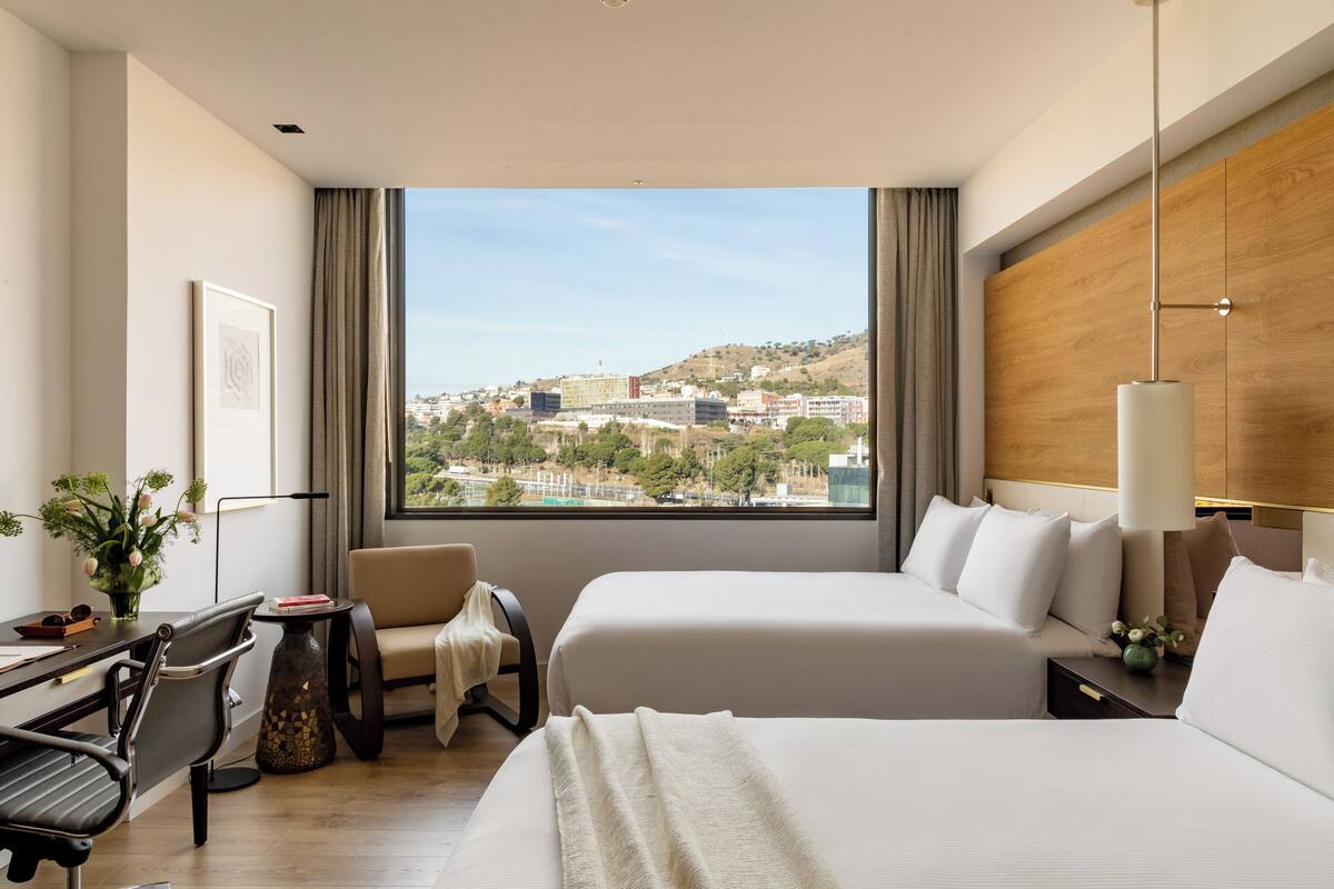 Habitación de hotel con dos camas, un escritorio, una silla y un ventanal que ofrece vista a la ciudad.