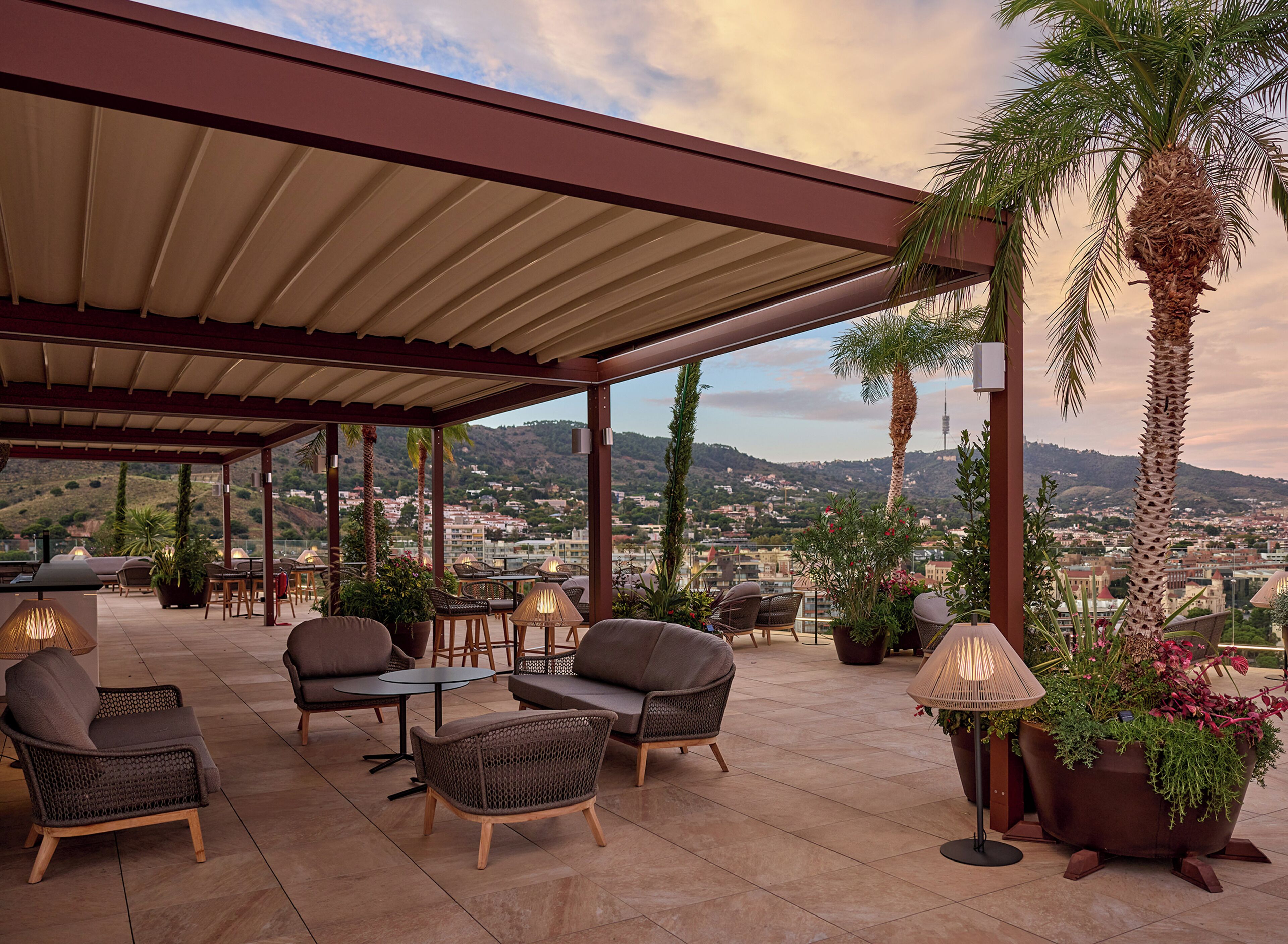 Un patio exterior con mobiliario de mimbre, techo cubierto y vistas a un paisaje urbano.