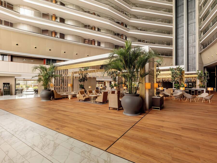 Un vestíbulo de hotel de varios niveles con una zona de estar central, plantas grandes en macetas y un techo alto.
