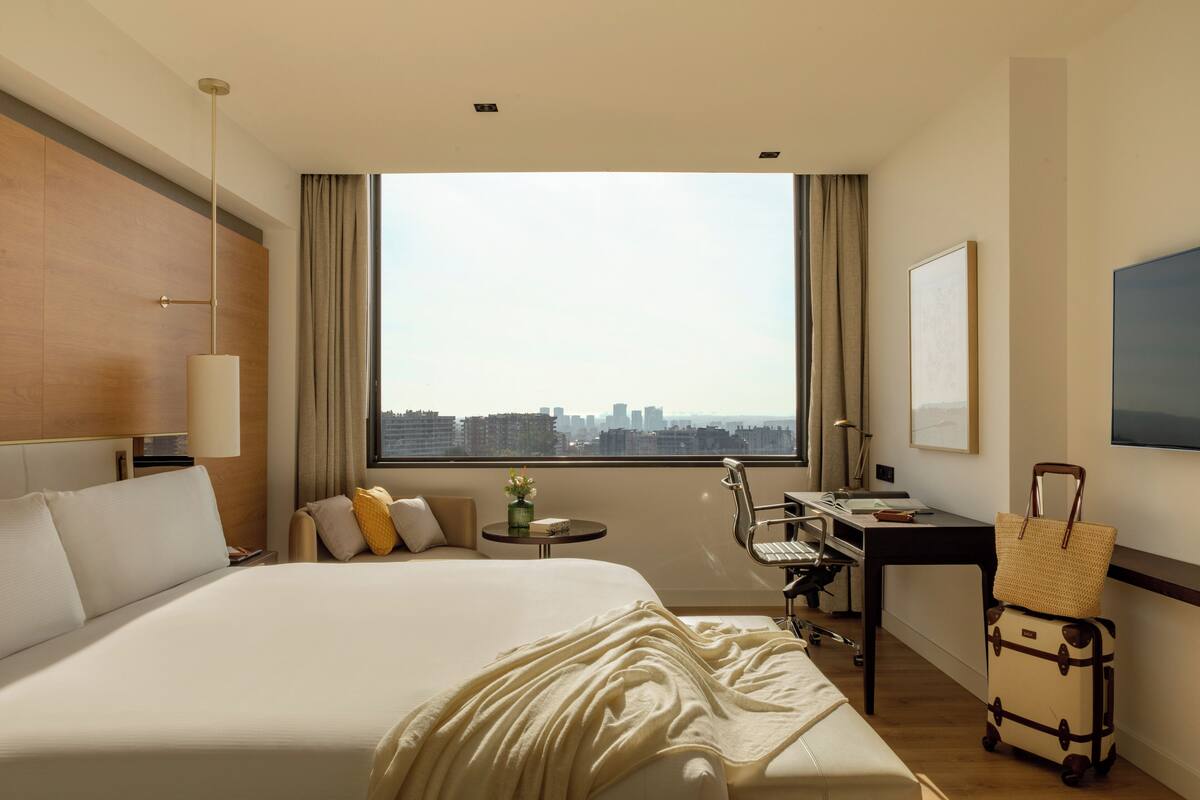 Habitación de hotel con una cama grande, un escritorio, una silla, una maleta y vistas a la ciudad.