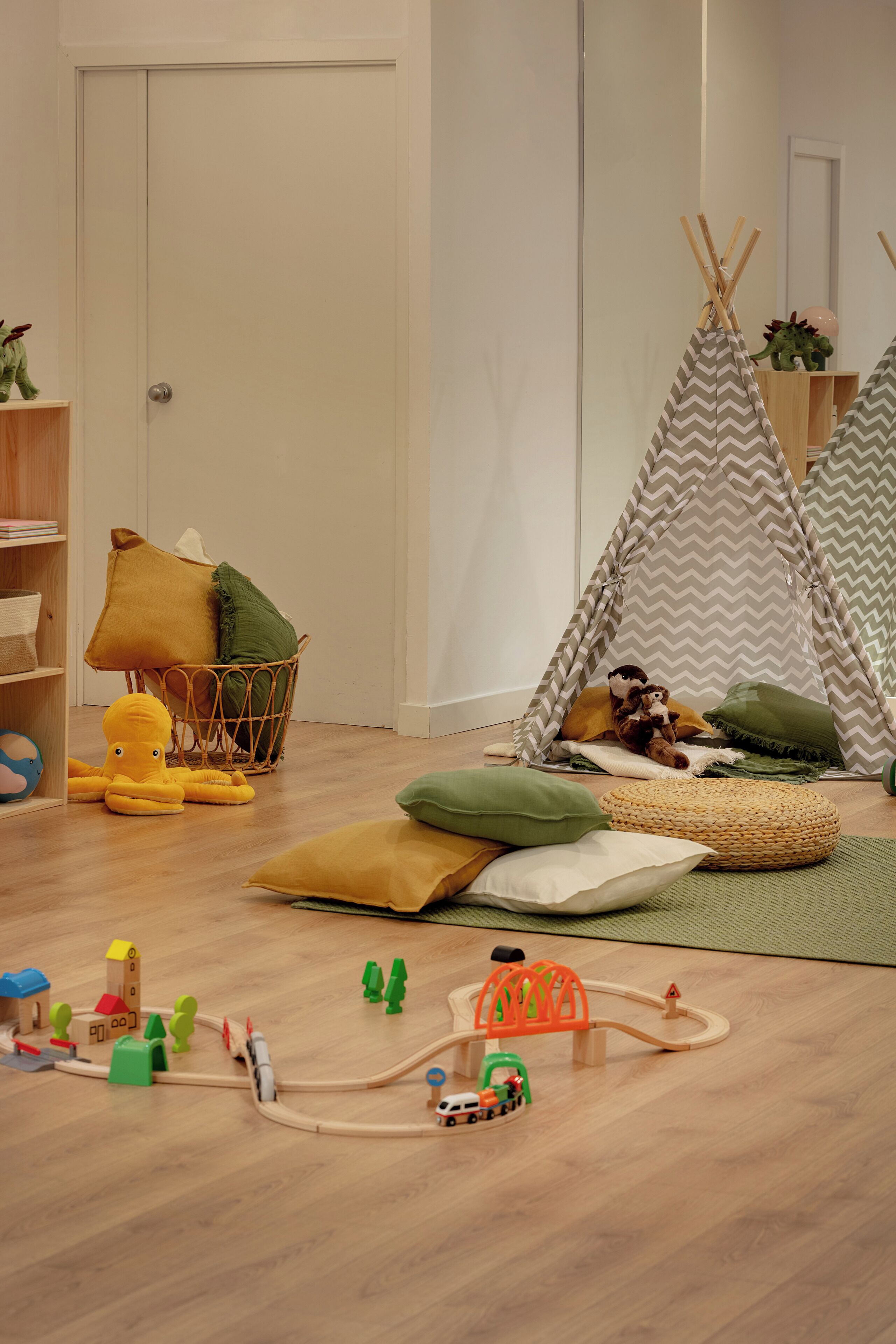 Una sala de juegos con piso de madera, una casita de juegos, un tipi y un juego de trenes de juguete.